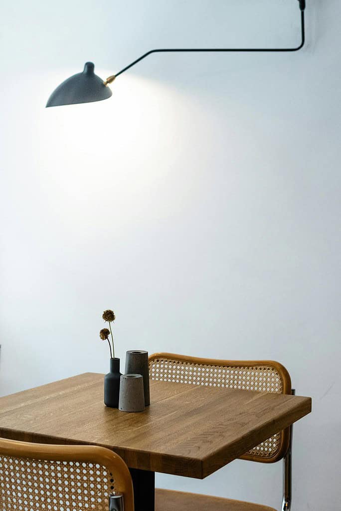 Square shaped wooden table with decorative flower and wicker chairs back and hanging lamp turned on near white wall in flat